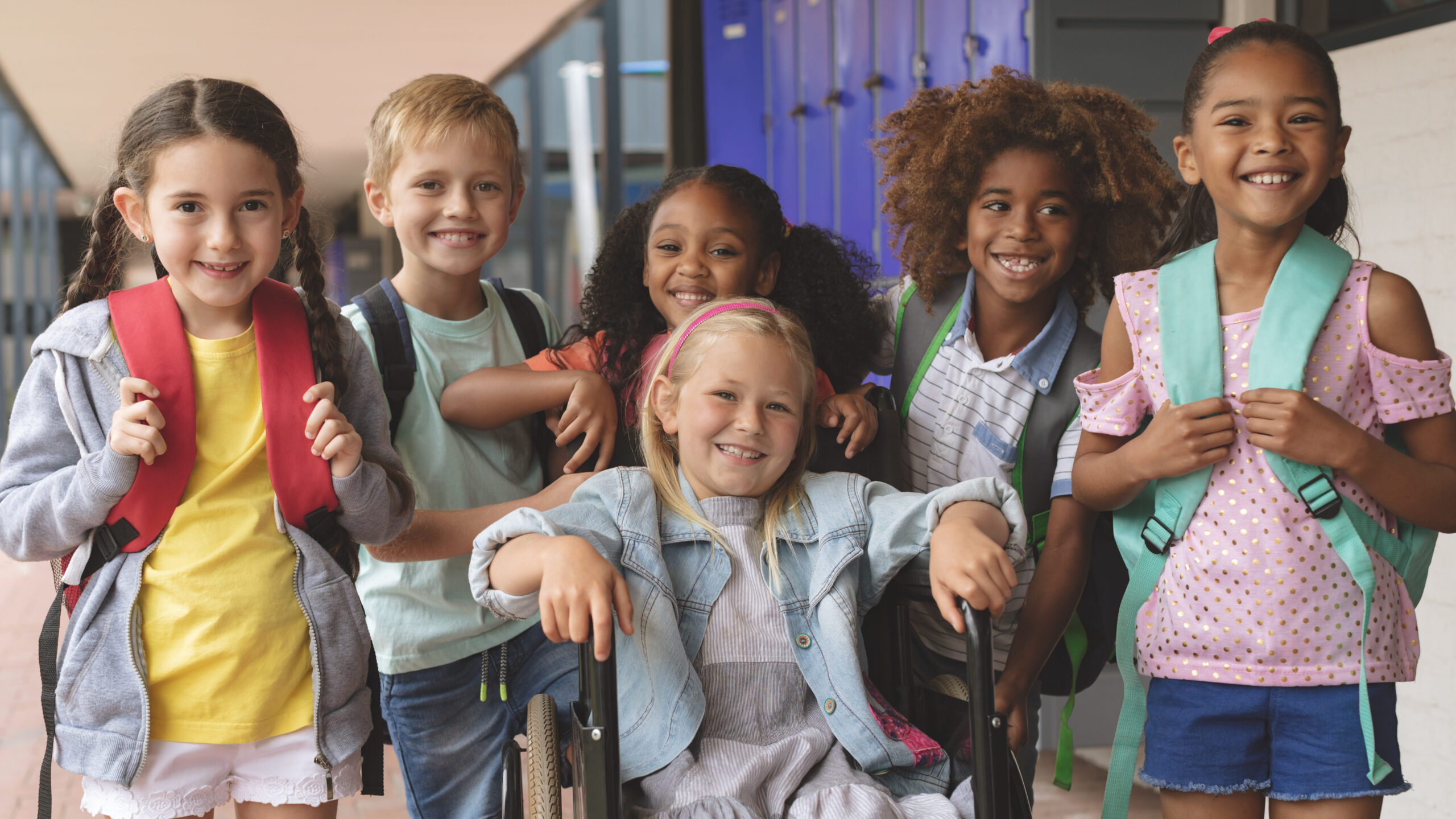 Front view of happy school kids standing in outside corridor at school while a Caucasian schoolgirl is sitting on wheelchair in foreground