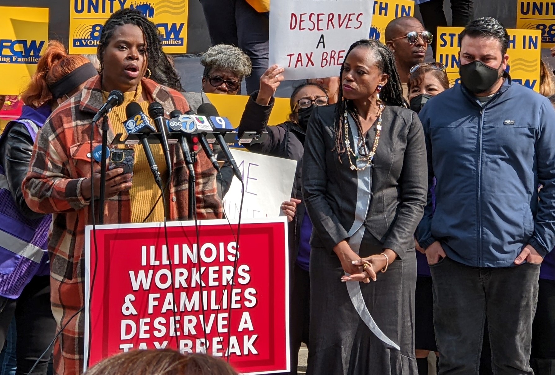 photo of ida speaking at podium in front of protesters with picket signs. Poster on podium reads 